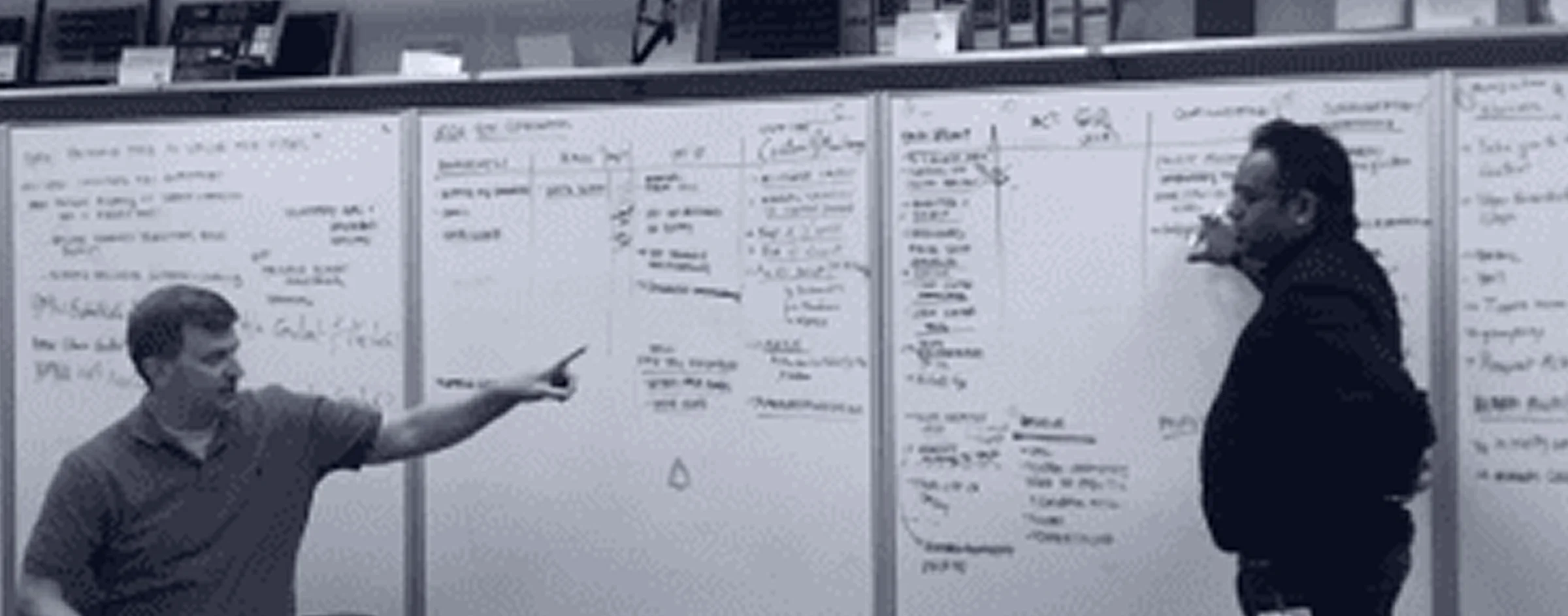 Two people at a whiteboard wall covered in notes, one pointing at it.